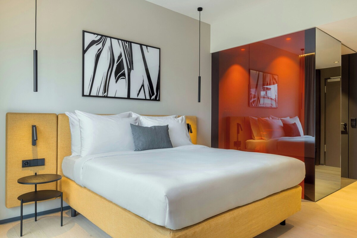 A modern hotel room with a large bed, a small side table, and a large abstract painting on the wall.