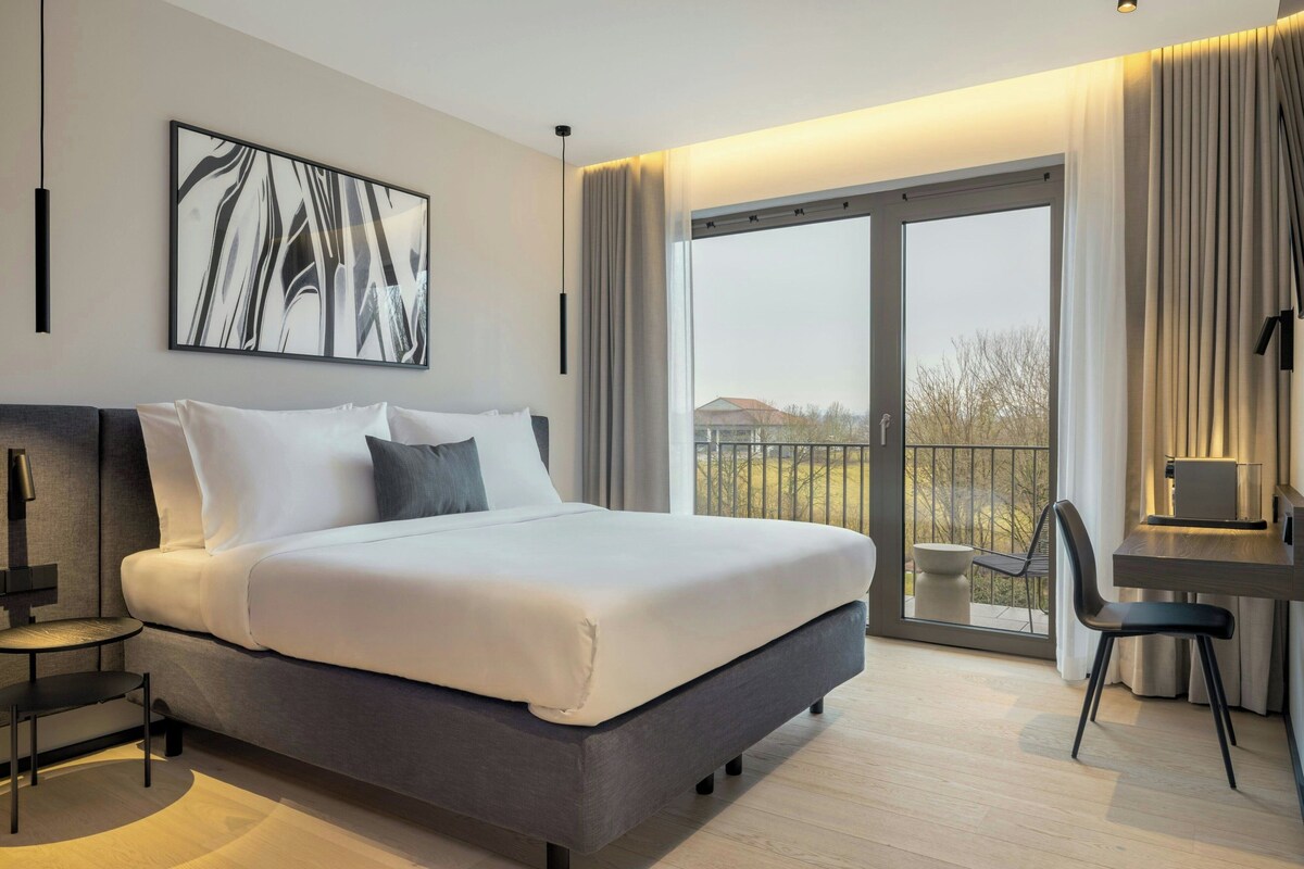 A modern hotel room with a large bed, a desk, and a view of the outdoors through a sliding glass door.