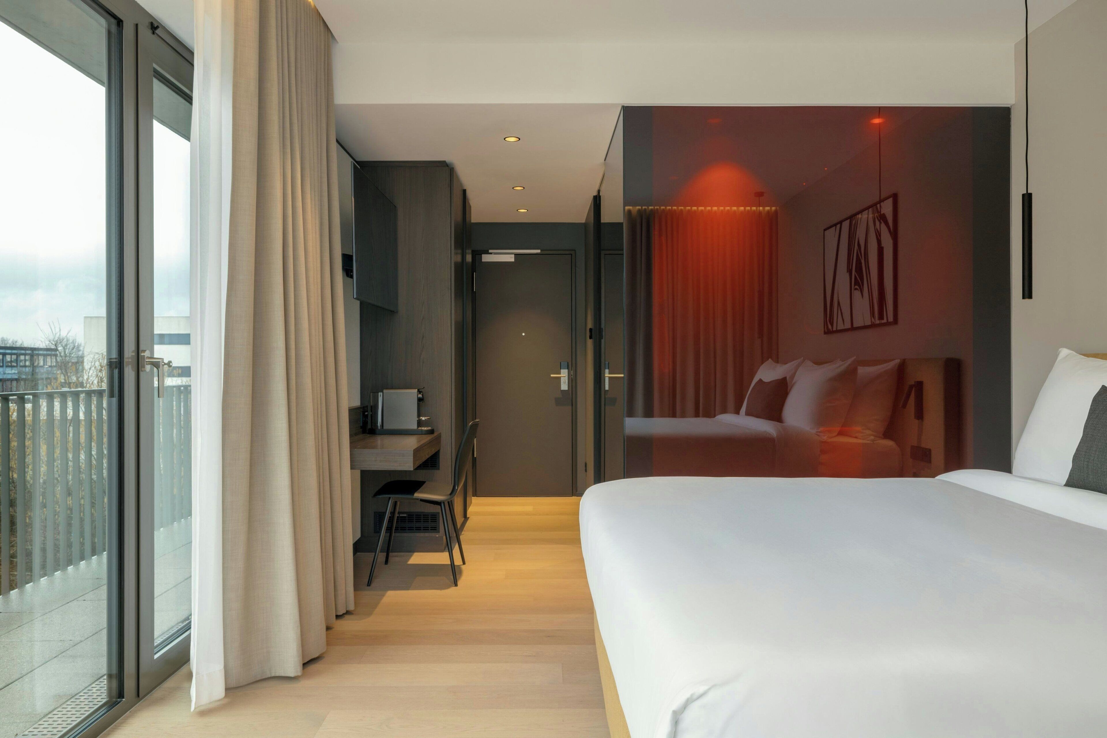 A modern hotel room with a large bed, a desk with a chair, a view through a sliding door, and a red accent wall.