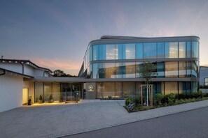 Un edificio moderno con fachada curva, amplios ventanales de vidrio y un área de entrada cuidada con plantas en macetas.
