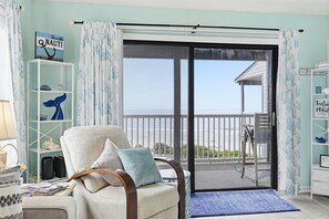 A room with a view of the ocean, a chair with cushions, a bookshelf, and a small table with a lamp.