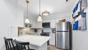 A modern kitchen with white cabinetry, a stainless steel refrigerator, a central island with bar stools, and pendant lighting. - Clean, Oceanfront Condo w/ Pool Access, King Suite, Bunks & Beautiful Views! (North Topsail Beach)
