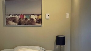 Una habitación de hotel con cama, mesita de noche, lámpara, un cuadro en la pared y un termostato.