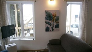 Una sala de estar con un sofá gris, un televisor de pantalla plana montado en la pared, un cuadro de hojas tropicales y una ventana con cortinas.