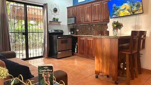 A compact kitchen with wooden cabinets, a microwave, and a coffee machine. There is a small island with stools and a TV mounted on the wall. - Poolside in Paradise: Walk to the Beach and More! (Coco)