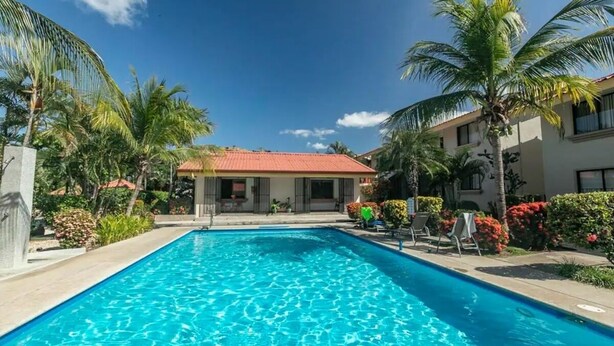 A swimming pool with clear blue water, surrounded by palm trees and a building with a red roof. - Poolside in Paradise: Walk to the Beach and More! (Coco)