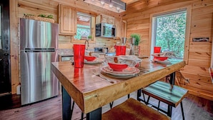 Une cuisine rustique aménagée dans une cabane en bois, avec un réfrigérateur en acier inoxydable, un four à micro-ondes et une table à manger en bois, agrémentée de bols et d’assiettes rouges.