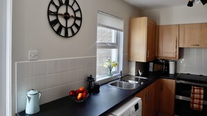 A kitchen with white tiles, wooden cabinets, a black countertop, a black clock, a window with a view, and a sink.