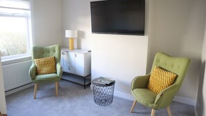 A room with two green armchairs, a small round side table, a flat-screen TV, a lamp, and a radiator.