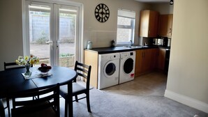 A kitchen with a washing machine, a dining table with chairs, and a clock on the wall.