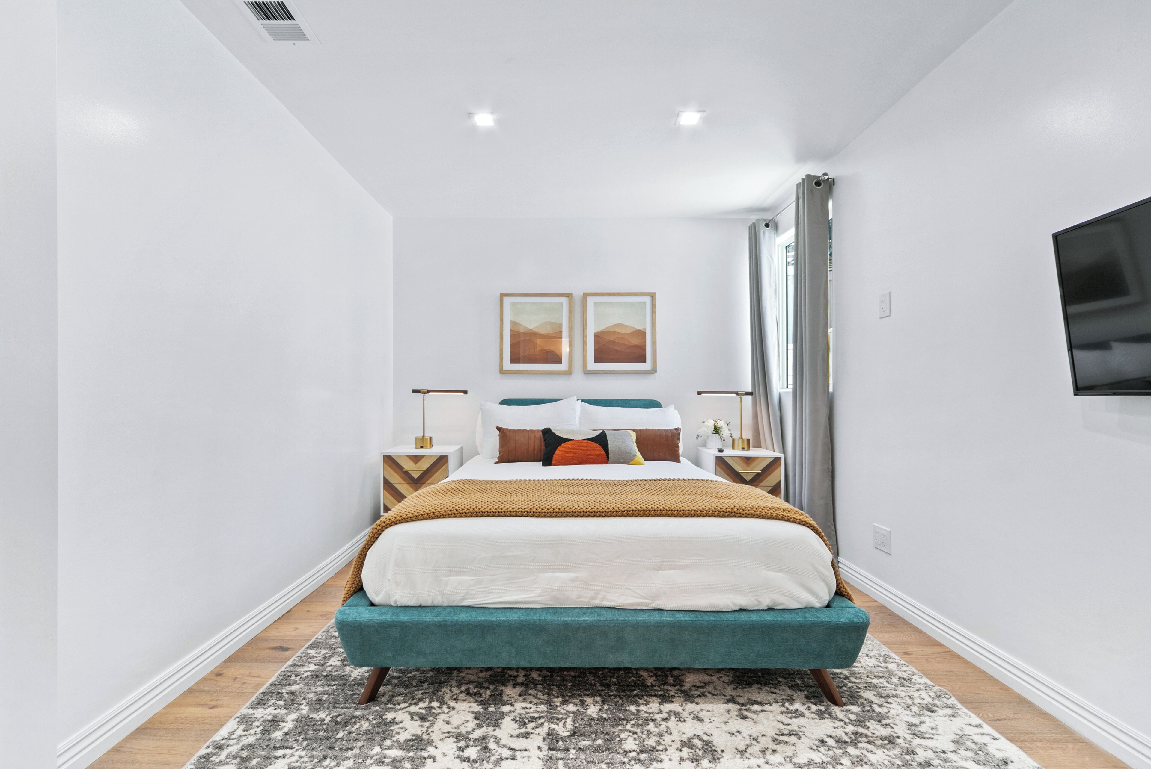 A modern bedroom with a teal bed, white walls, and a large window with curtains.