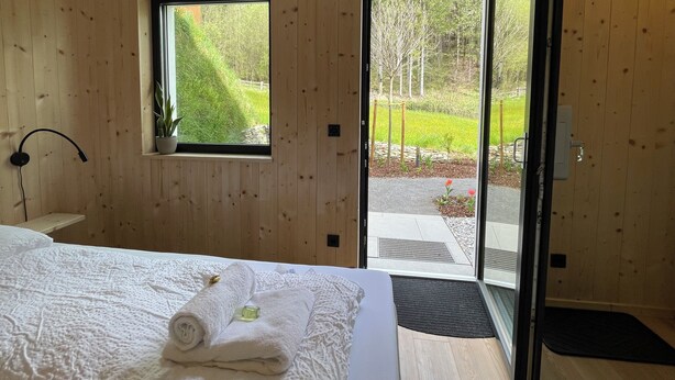 A modern bedroom with a wooden interior, a large bed with white linens, a potted plant, and a view of a garden through a glass door. - Cozy Double Room - Private Entrance – Easy Self Check-In (Rum)