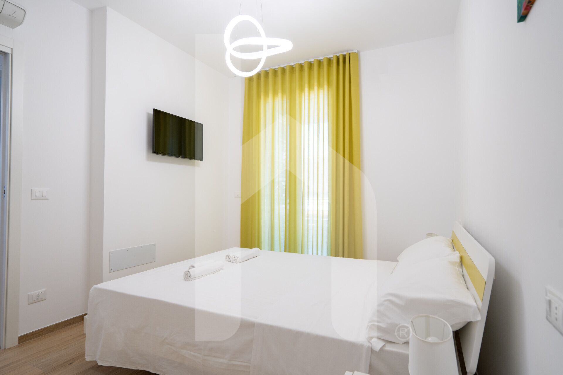 A modern hotel room with a bed, a flat-screen TV, and a window with yellow curtains.