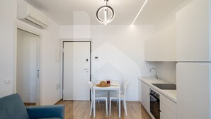 Una cocina moderna con armarios blancos, horno empotrado y un lavamanos. Hay una zona de comedor con una mesa y sillas, y un sofá azul en la sala de estar.