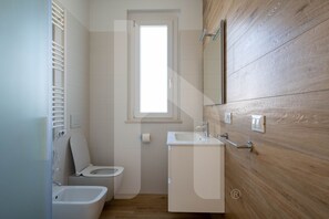 Une petite salle de bain moderne, équipée de toilettes, d’un bidet et d’un lavabo. Une fenêtre se trouve au-dessus des toilettes et un panneau en bois orne le mur de droite.