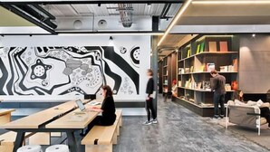 Une bibliothèque moderne dotée d’une grande fresque murale abstraite, de tables en bois et d’étagères à livres.