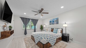 Un dormitorio con cama, ventilador de techo, televisión montada en la pared, mesita de noche con lámpara y ventana con cortinas.