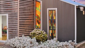 A modern building with a wooden slatted facade, a colorful stained glass window, and a small garden with white flowers. - Cozy cabin in peaceful Wangoom with AC, WiFi (Wangoom)