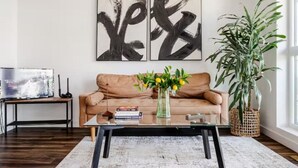 Ein Wohnzimmer mit einem braunen Ledersofa, einem Glastisch, einer Vase mit gelben Blumen und einer großen Grünpflanze.