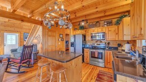 A wooden cabin interior with a kitchen featuring a central island, bar stools, a refrigerator, stove, and a hanging pot rack. - Magic’s Rest 2 Bed Cabin located in Peaceful Gated Community with lake access (Lake Lure)
