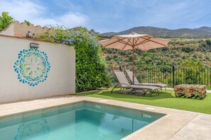 Una piscina con solárium adoquinado, una silla de descanso, una sombrilla y vistas a un paisaje montañoso.