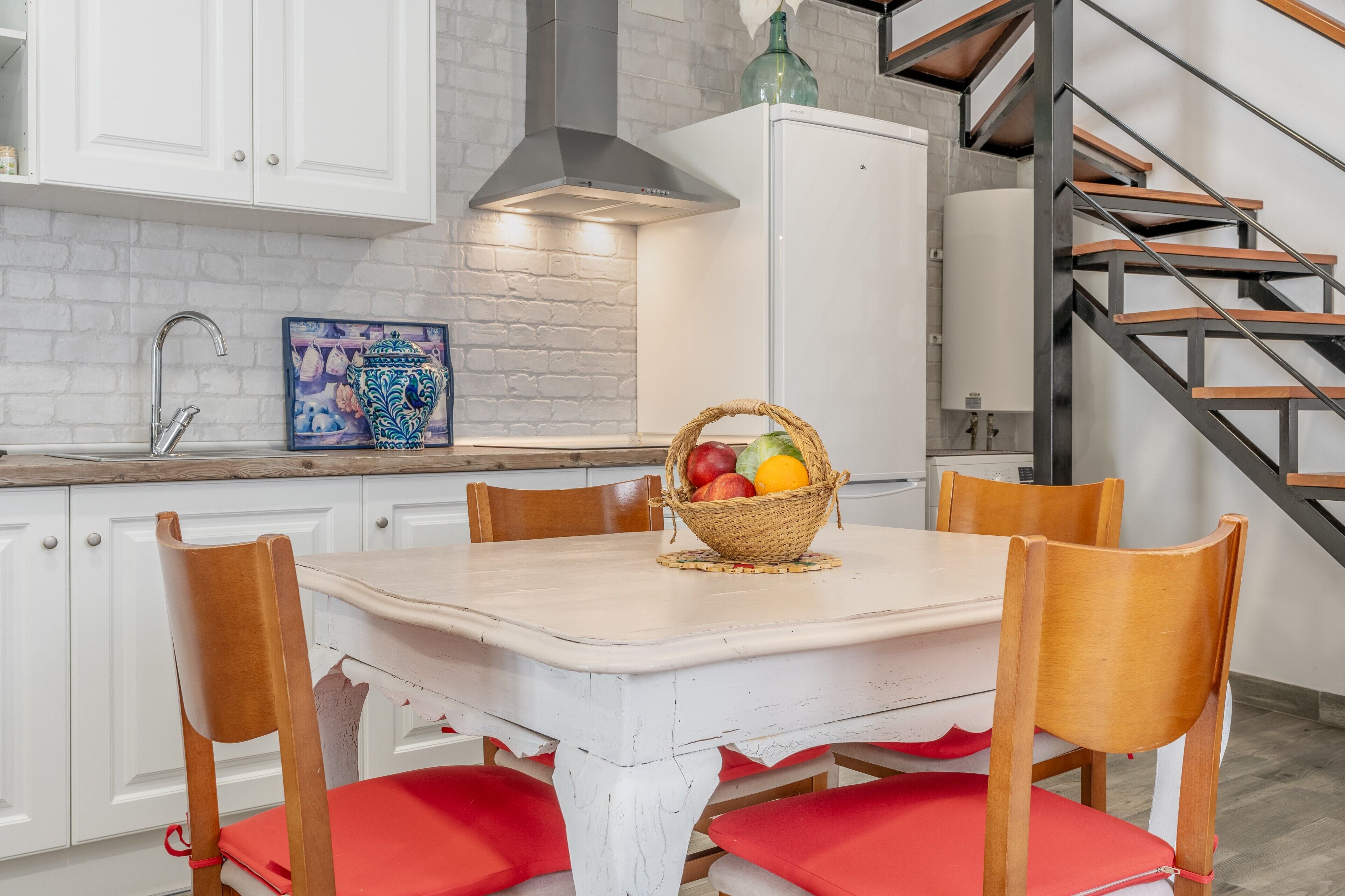 A kitchen with white cabinets, a white table, red cushioned chairs, a wicker fruit basket, and a staircase with wooden steps.
