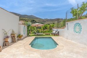 A swimming pool with a sun lounger and a small deck area, surrounded by potted plants and a view of hills.