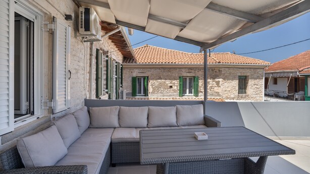 A covered outdoor seating area with a sofa and table, stone buildings in the background, and a clear sky. - Holiday Home 'Sparto' with Private Terrace, Wi-Fi and Air Conditioning (Spartochori)