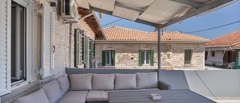 A covered outdoor seating area with a sofa and table, stone buildings in the background, and a clear sky.