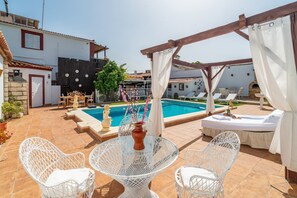 Un área junto a la piscina con mesa de vidrio, sillas de ratán blanco, un espacio de descanso cubierto y una piscina.