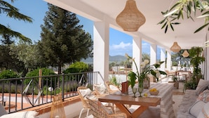 Eine sonnendurchflutete Terrassengrundstück mit Korbmöbeln, einem Holztisch und Blick auf Bäume und Berge.