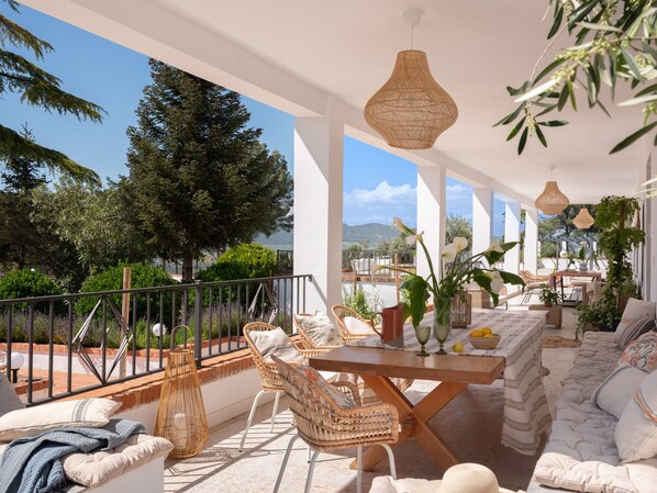 A sunlit outdoor patio with wicker furniture, a wooden table, and a view of trees and mountains.