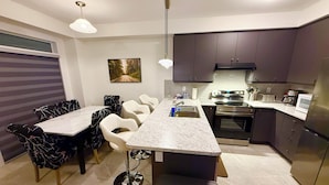 A modern kitchen with dark cabinets, a marble countertop, and stainless steel appliances. There is a dining area with a table and chairs.