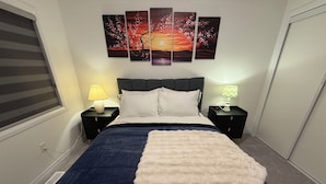 A bedroom with a bed, two bedside tables, a nightstand, a wall-mounted lamp, and a window with blinds.