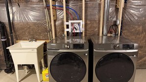 Two Samsung front-loading washing machines, a white sink, and visible plumbing and electrical wiring in a room under construction.