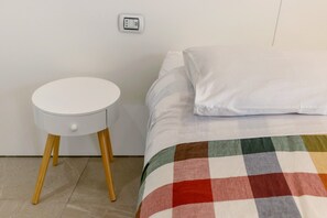 Un lit avec une couverture colorée, un oreiller blanc et une petite table de chevet blanche dotée d’un tiroir.