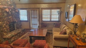 A cozy living room with a stone fireplace, a wooden coffee table, patterned sofas, and a lamp. - 3 BRD Apartment (Bigfork)