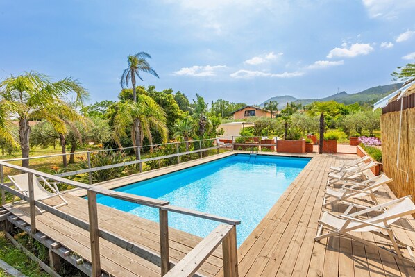 A wooden deck with a pool, lounge chairs, and a view of a house and mountains.