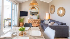A modern living room with a grey sofa, a dining table with chairs, a TV, and a kitchen area visible through a glass door. - Apartment 'La Terrasse Du Golf' with Shared Pool, Private Terrace and Balcony (Talmont-Saint-Hilaire)