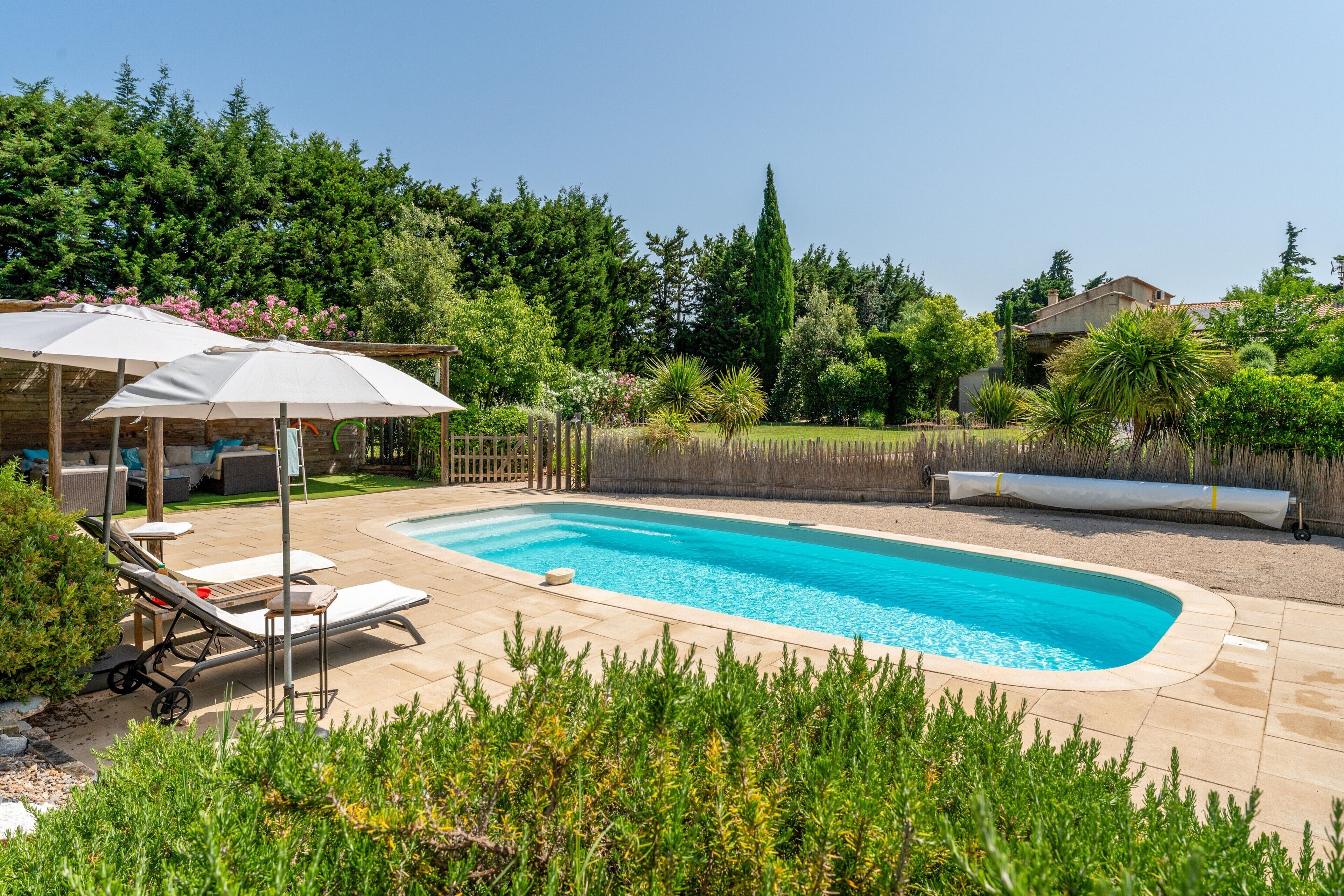 A swimming pool with lounge chairs and umbrellas, surrounded by greenery and a wooden fence.