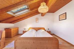Une chambre avec un plafond en bois, une lucarne, un lit avec du linge de lit blanc, une commode en bois et une table de chevet avec une lampe.