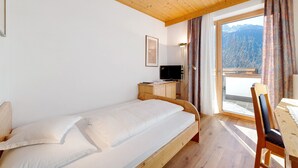 A single bed with a wooden headboard, a bedside table with a television, and a view of mountains through a large window.