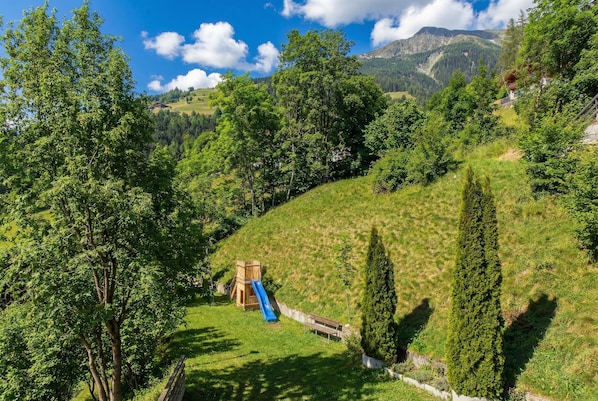 Une aire de jeux avec un toboggan et une structure d’escalade, entourée d’arbres et bordée d’une colline.