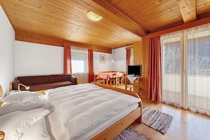 Træloft, seng med hvide sengetøj, sofa, fjernsyn, spisebord og et vindue med gardiner.