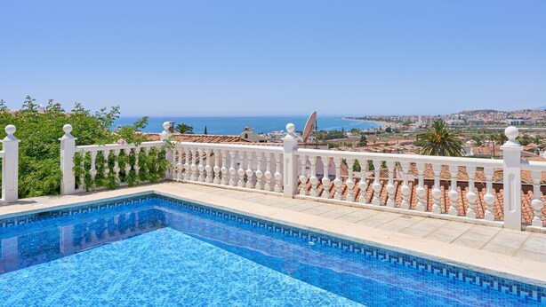 A swimming pool with a white balustrade, a clear view of the sea, and a cityscape in the background.