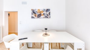 Una mesa de comedor blanca con un control remoto, una lámpara de mesa tejida y una obra de arte con motivos de hojas en la pared.