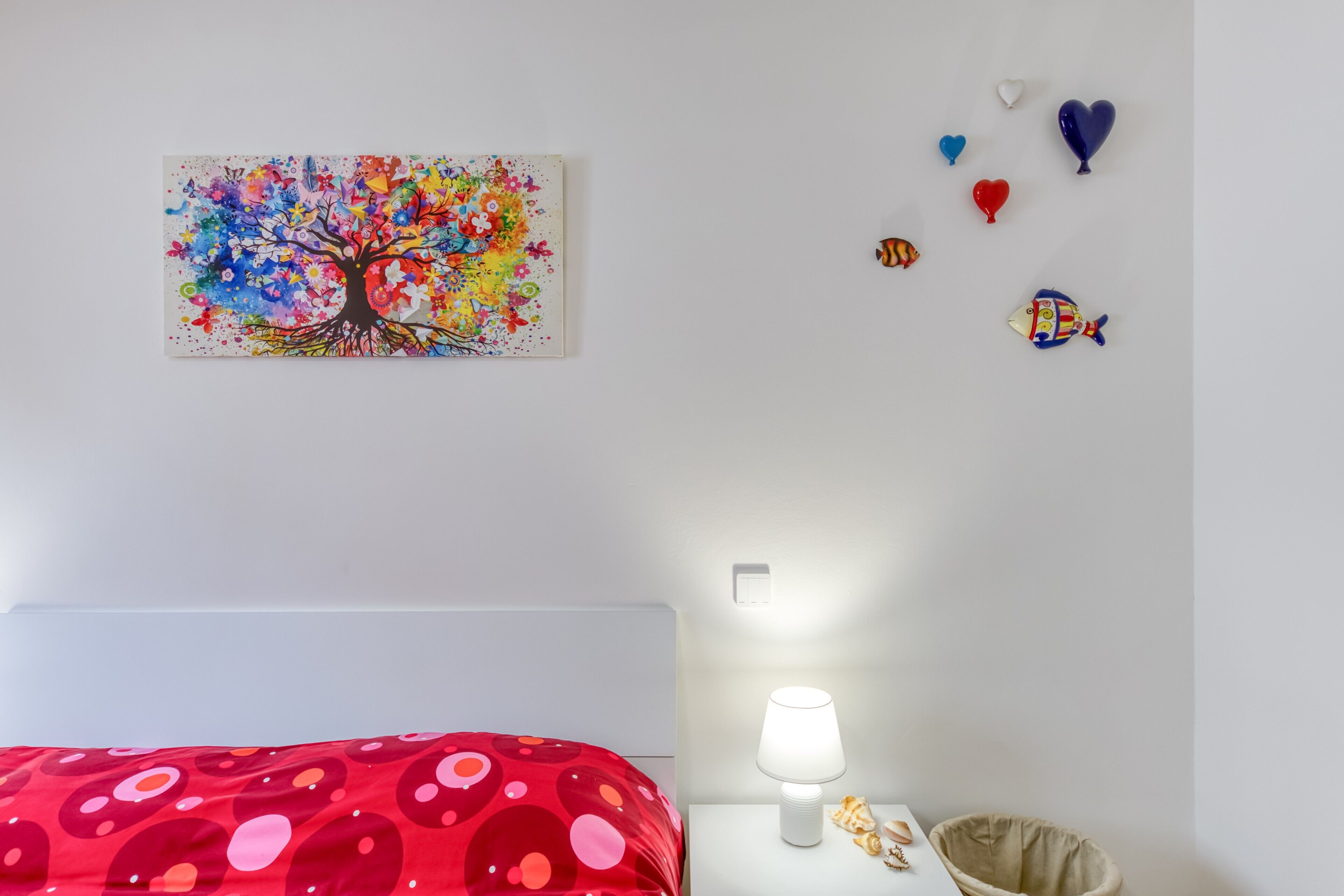 Een slaapkamer met een rood bed met stippen, een kleurrijk schilderij van een boom aan de muur, een nachtkastje met een lamp, en decoratieve vissen en hartvormige ballonnen.