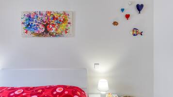 Een slaapkamer met een rood bed met stippen, een kleurrijk schilderij van een boom aan de muur, een nachtkastje met een lamp, en decoratieve vissen en hartvormige ballonnen.
