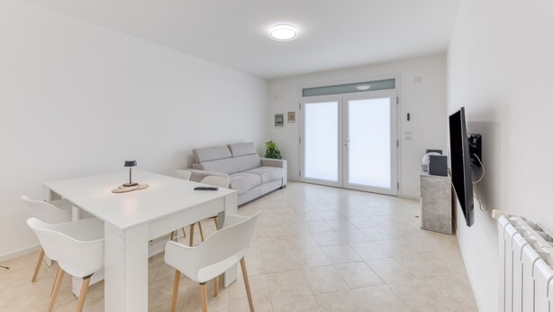 Una sala de estar moderna con un sofá blanco, una mesa de comedor con sillas, un televisor montado en la pared y un amplio ventanal corredizo que da a un balcón.