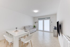 Ein modernes Wohnzimmer mit einem weißen Sofa, einem Esstisch mit Stühlen, einem an der Wand montierten Fernseher und einer Glastür zum Balkon.